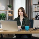 Beautiful caucasian woman with long dark hair sitting at desk and typing on laptop.